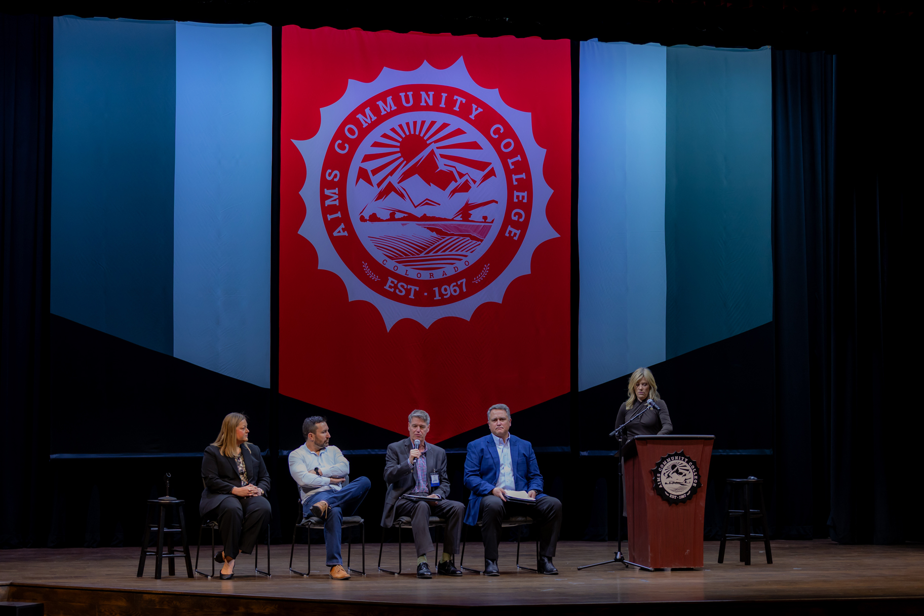A panel of four speakers and a moderator at the podium on the Aims Community College stage at the 2025 Confluence and Net Zero Cities
