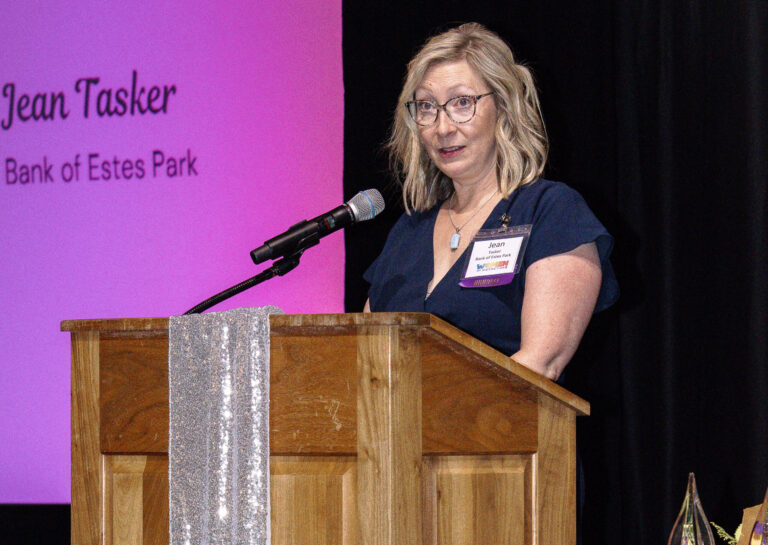 A speaker at the podium below a screen reading "Jean Tasker, Bank of Estes Park"