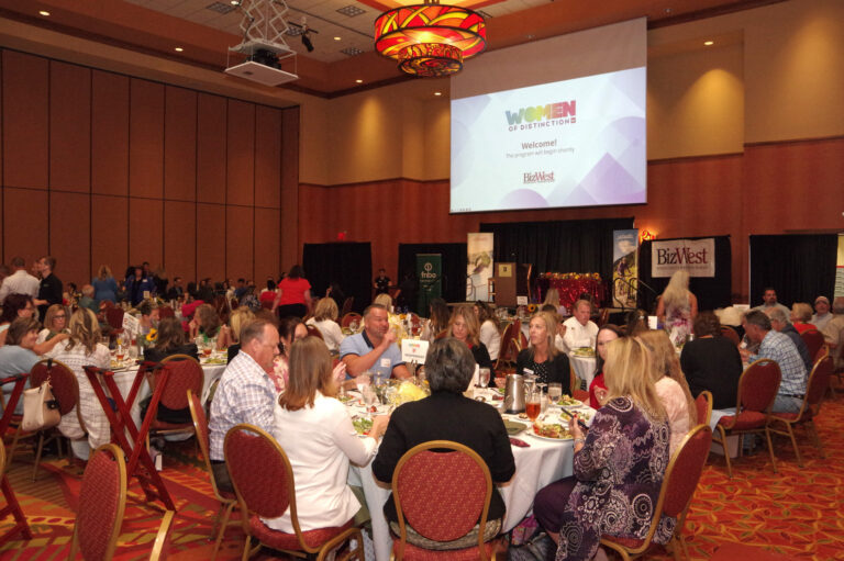 The ballroom filled with guests at round tables facing the stage screen