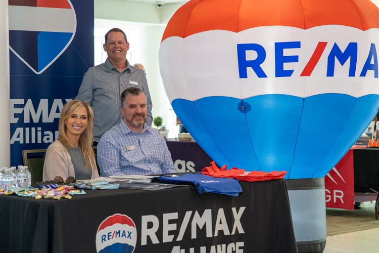 Three people at the RE/MAX Alliance sponsor table beside an inflatable RE/MAX hot-air balloon