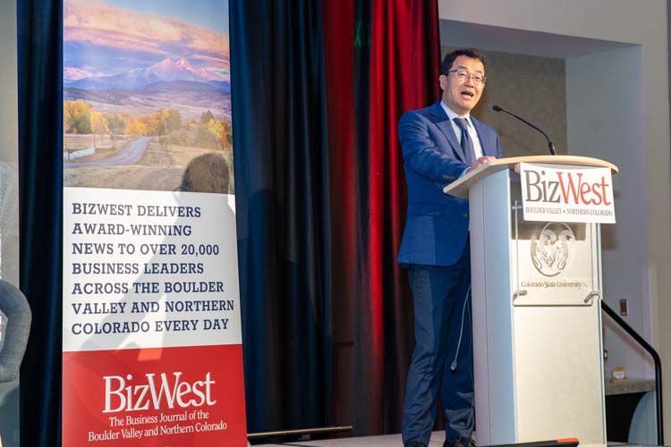 A speaker in a blue suit delivers remarks from the BizWest lectern beside a BizWest banner