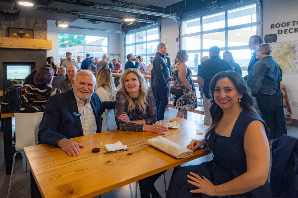 Three guests seated at a wooden table, smiling, with a crowd mingling behind them at a BizWest reception