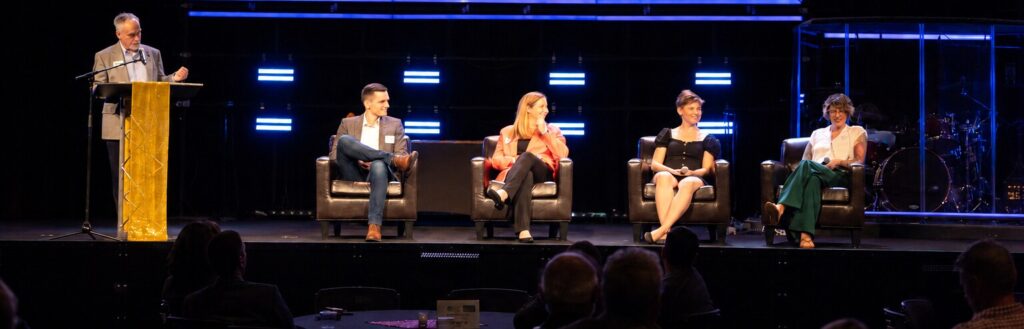A moderator at a podium and four panelists seated in armchairs on a stage with blue lighting at a BizWest event