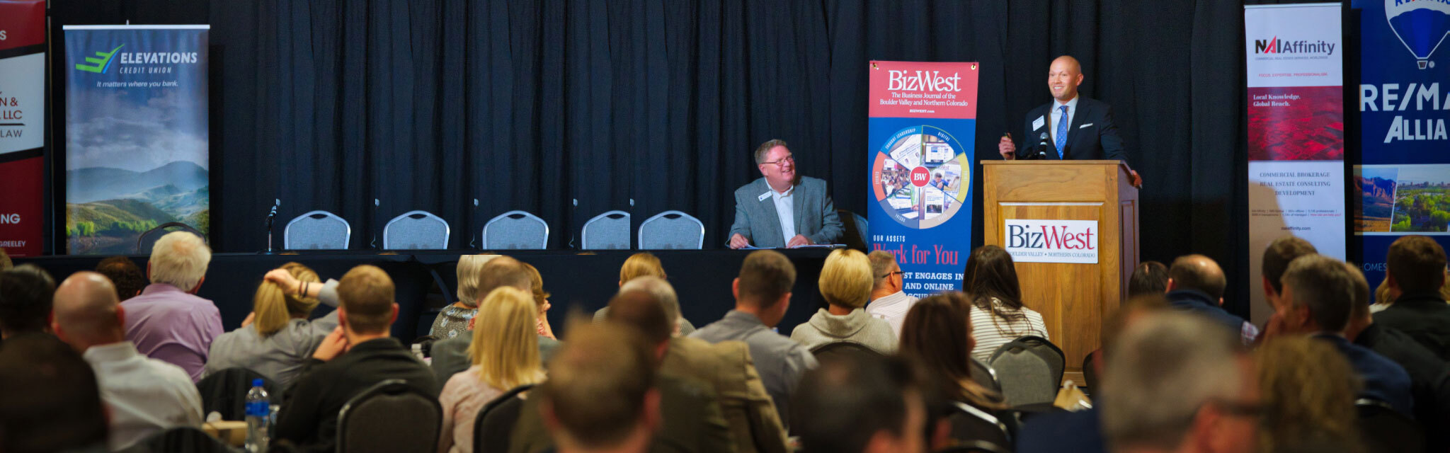 A speaker addresses the audience from the BizWest podium at the Northern Colorado Real Estate Summit, with a second panelist seated at the head table
